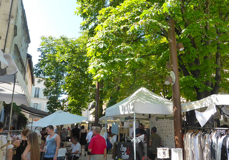 Les étals du marché dans une rue ombragée d'arbres plantés, bordée de hautes maisons en pierre aux volets, Saint-Remy Provence